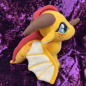 May include: A plush yellow dragon with brown horns, red wings, and green eyes. The dragon has a white belly and a white stripe down its back.