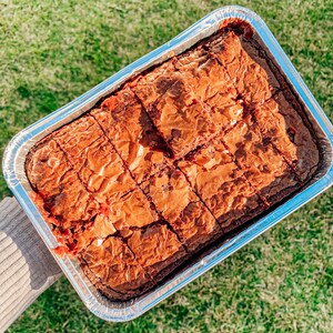 May include: A foil tray filled with freshly baked brownies, cut into squares, rests on green grass. The brownies are a deep brown with a cracked surface. The foil tray is silver. The image suggests a homemade treat, perfect for sharing.