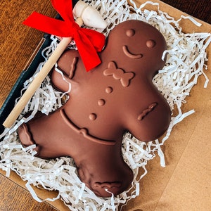 May include: A large, dark chocolate gingerbread man in a cardboard box with white paper filler. A wooden stirring stick with a red bow and a white chocolate piece are also in the box. The gingerbread man has a smiling face and bow tie.