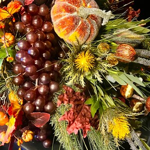 Puede incluir: Una corona decorativa oto&ntilde;al con una calabaza, uvas y follaje. La calabaza es naranja y amarilla con purpurina. Las uvas son de color p&uacute;rpura oscuro, y las hojas son rojas, naranjas y verdes, creando un ambiente festivo.