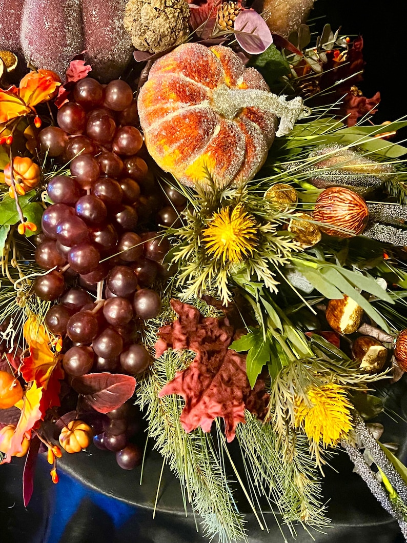 Puede incluir: Una corona decorativa oto&ntilde;al con una calabaza, uvas y follaje. La calabaza es naranja y amarilla con purpurina. Las uvas son de color p&uacute;rpura oscuro, y las hojas son rojas, naranjas y verdes, creando un ambiente festivo.