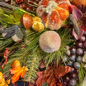 Puede incluir: Primer plano de un arreglo decorativo con tem&aacute;tica oto&ntilde;al. Presenta una calabaza naranja azucarada, uvas y follaje oto&ntilde;al. El arreglo incluye agujas de pino, bayas y hojas en tonos naranja, rojo y verde. El tema general es el oto&ntilde;o y la cosecha.