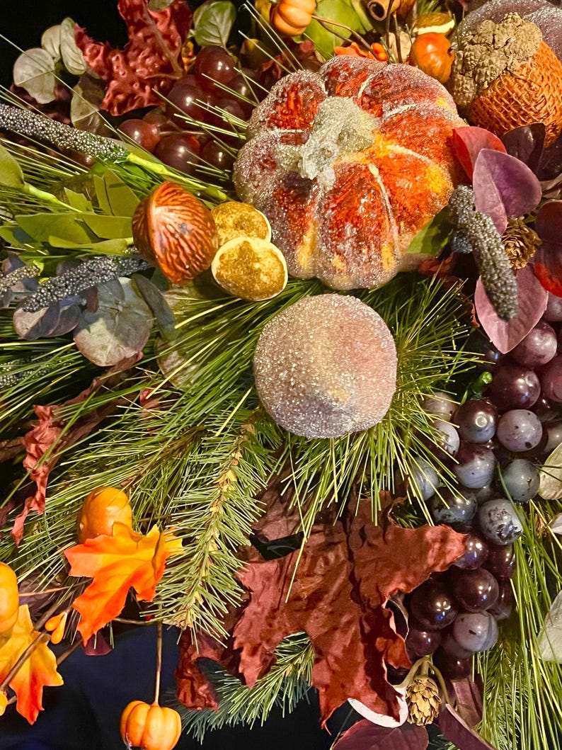 Puede incluir: Primer plano de un arreglo decorativo con tem&aacute;tica oto&ntilde;al. Presenta una calabaza naranja azucarada, uvas y follaje oto&ntilde;al. El arreglo incluye agujas de pino, bayas y hojas en tonos naranja, rojo y verde. El tema general es el oto&ntilde;o y la cosecha.