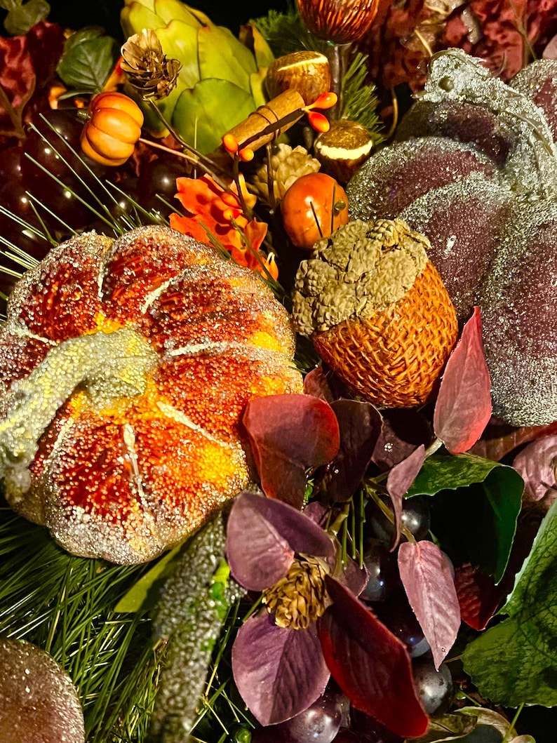 Puede incluir: Primer plano de un arreglo oto&ntilde;al con una calabaza grande y naranja con purpurina, una bellota y varias hojas. El arreglo incluye peque&ntilde;as calabazas, bayas y vegetaci&oacute;n, creando una exhibici&oacute;n festiva.
