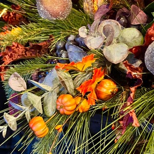 Puede incluir: Primer plano de un arreglo floral con tem&aacute;tica oto&ntilde;al. El arreglo presenta calabazas naranjas, agujas de pino verdes y hojas coloridas. Los elementos decorativos incluyen orbes marrones esmerilados y uvas moradas. La paleta de colores es oto&ntilde;al.