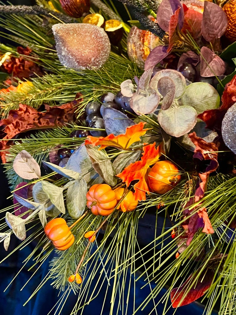 Puede incluir: Primer plano de un arreglo floral con tem&aacute;tica oto&ntilde;al. El arreglo presenta calabazas naranjas, agujas de pino verdes y hojas coloridas. Los elementos decorativos incluyen orbes marrones esmerilados y uvas moradas. La paleta de colores es oto&ntilde;al.
