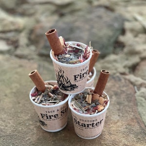 Könnte beinhalten: Drei kleine Becher gefüllt mit Feueranzündermaterial, darunter Zedernholz, Kräuter und Botanicals. Jeder Becher hat einen Zimtstange obenauf und ein Etikett mit der Aufschrift "Fire Starter".