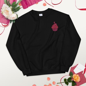 May include: Black sweatshirt featuring a small, embroidered cupcake design in pink hues. The cupcake is adorned with hearts. The sweatshirt is made of a soft material, suitable for casual wear.