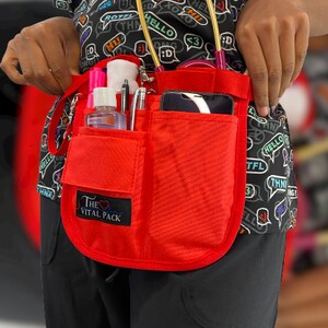 May include: Red medical utility belt with multiple pockets and a phone holder. The belt has a black patch with the text "The Vital Pack" and a heart symbol.