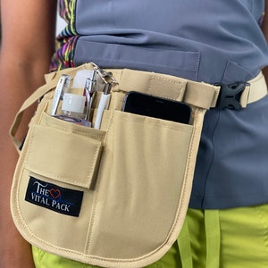 May include: A beige canvas belt with multiple pockets and a black buckle. The belt has a small patch with the text "The Vital Pack" and a red heart symbol. The belt is worn over a gray shirt and green pants.