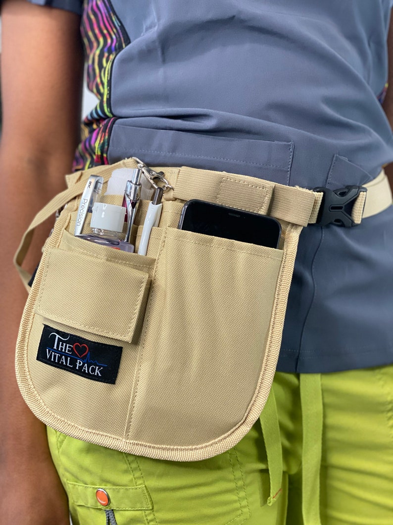 May include: A beige canvas belt with multiple pockets and a black buckle. The belt has a small patch with the text "The Vital Pack" and a red heart symbol. The belt is worn over a gray shirt and green pants.