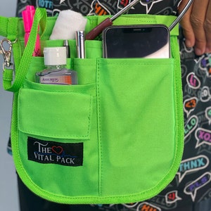 May include: A green medical utility belt with multiple pockets, a cell phone, a stethoscope, a bottle of hand sanitizer, and a pen. The belt has a black label with the text "The Vital Pack" and a red heart with a heartbeat graphic.