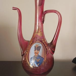 Op de afbeelding: Een robijnrode glazen karaf met een lange tuit en handvat. Het heeft een portret van een man in militair uniform. Gouden accenten sieren het oppervlak van de karaf en het heeft een decoratieve stop.