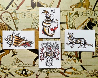 May include: Four rectangular art prints featuring traditional tattoo-style illustrations: a conch shell, a sea goat, a cat, and a bull head surrounded by flames and smoke. Each print is white with black outlines and vibrant color accents.
