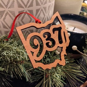 May include: A wooden ornament in the shape of Ohio with the numbers "937" cut out. The ornament is light brown and has a red ribbon for hanging. It rests on a bed of greenery and white berries.