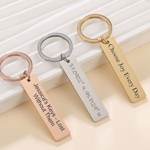 May include: Three silver, gold, and rose gold keychains with engraved tags. The silver tag reads "37.0902° N, 95.7129° W". The gold tag reads "Choose Joy Every Day". The rose gold tag reads "Jessica's Keys - Lost Without Them".