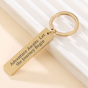 Può includere: Portachiavi dorato con un'etichetta rettangolare su cui è scritto "Adventure Awaits Let the Journey Begin".