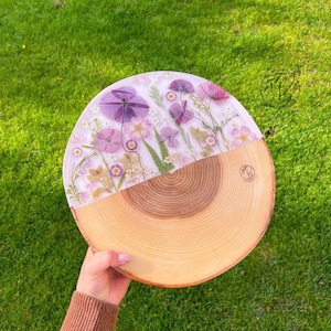 May include: A round wooden serving tray with a clear epoxy resin top featuring pressed purple and white flowers. The tray is made from a natural wood slice with visible growth rings.