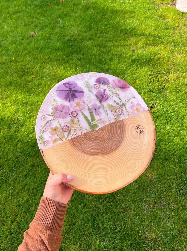 May include: A round wooden serving tray with a clear epoxy resin top featuring pressed purple and white flowers. The tray is made from a natural wood slice with visible growth rings.
