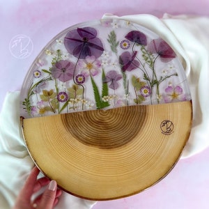 May include: A round wooden serving tray with a clear resin top featuring pressed purple and white flowers. The tray is half wood and half resin, with a natural wood grain pattern on the wood side. The resin side features a variety of pressed flowers, including purple poppies, white daisies, and green leaves.