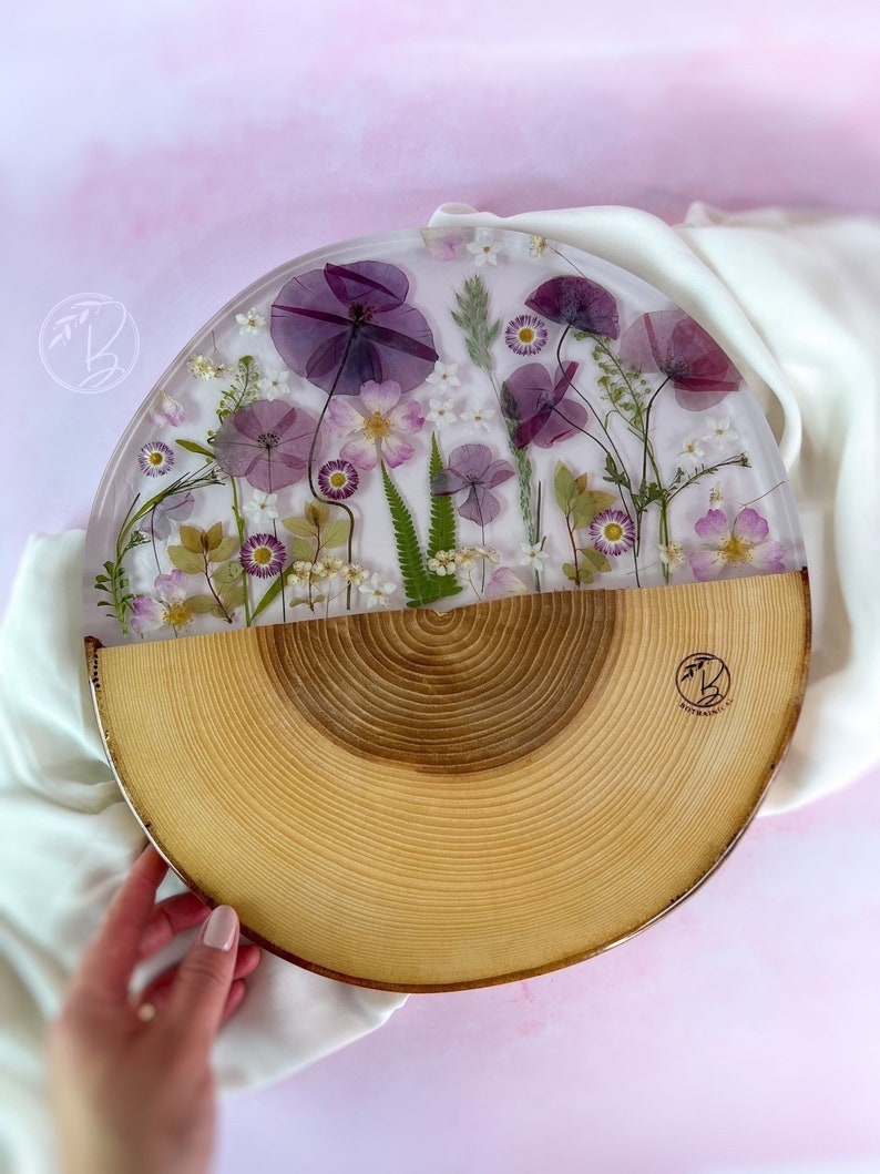 May include: A round wooden serving tray with a clear resin top featuring pressed purple and white flowers. The tray is half wood and half resin, with a natural wood grain pattern on the wood side. The resin side features a variety of pressed flowers, including purple poppies, white daisies, and green leaves.