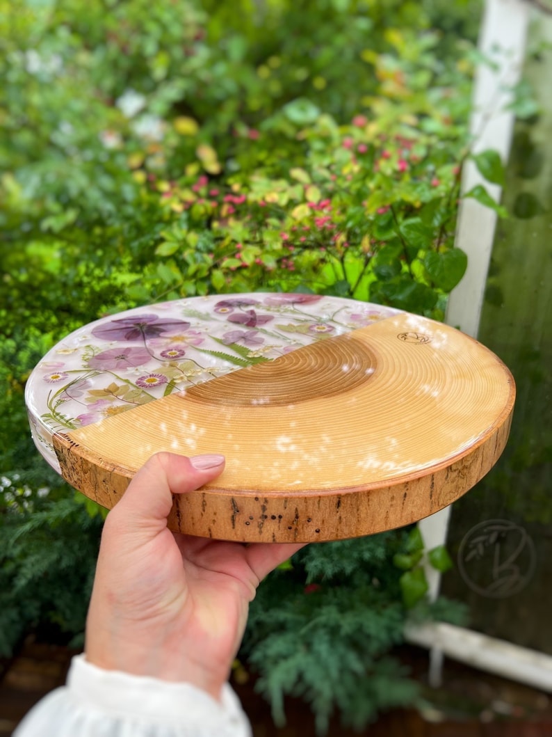 May include: A round wooden coaster with a clear resin top featuring pressed flowers in shades of purple and white. The coaster has a natural wood bark edge.