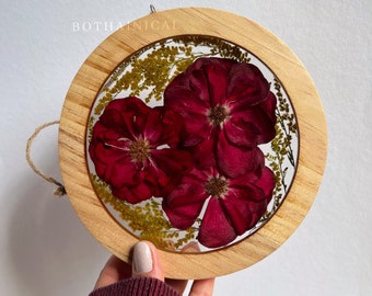 May include: A round wooden frame with a clear resin insert containing pressed red roses and yellow flowers. The frame has a small metal loop for hanging.