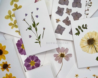 May include: A set of ten blank greeting cards featuring pressed flowers. The cards are white with a variety of flowers, including yellow, purple, pink, and blue.
