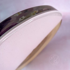 May include: A round wooden coaster with a pink resin top. The resin is clear and has embedded dried flowers. The coaster is perfect for protecting surfaces from scratches and spills.