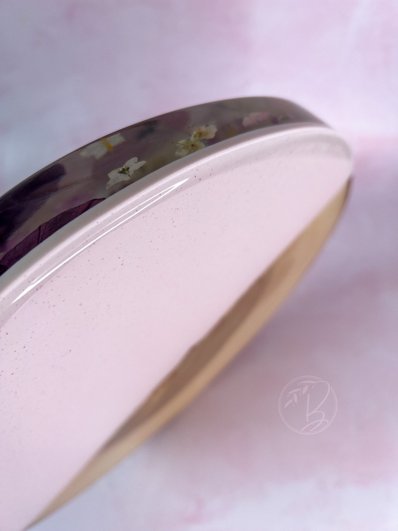 May include: A round wooden coaster with a pink resin top. The resin is clear and has embedded dried flowers. The coaster is perfect for protecting surfaces from scratches and spills.