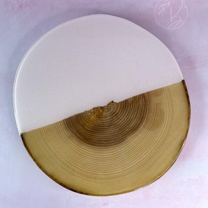 May include: A round wooden serving tray with a light pink epoxy resin top. The tray has a natural wood finish and a unique wood grain pattern.