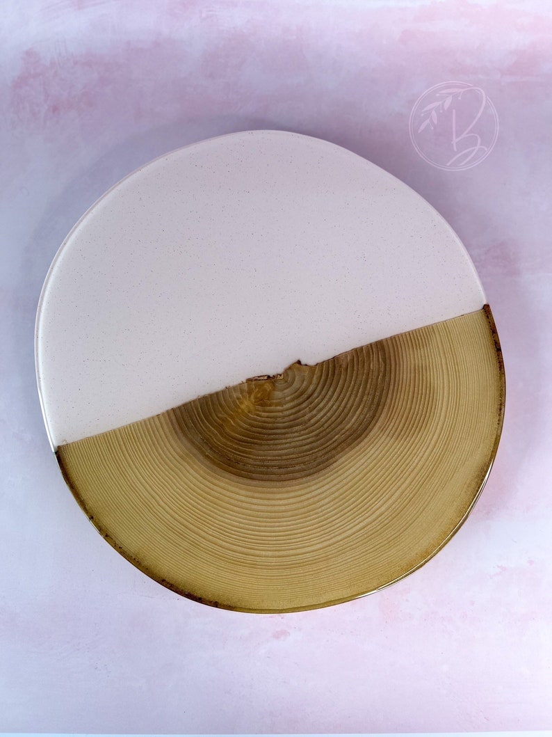 May include: A round wooden serving tray with a light pink epoxy resin top. The tray has a natural wood finish and a unique wood grain pattern.