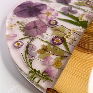 May include: A round, clear resin coaster with pressed flowers and leaves. The coaster has a light pink background and features purple, white, and green flowers. The coaster is attached to a wooden base.