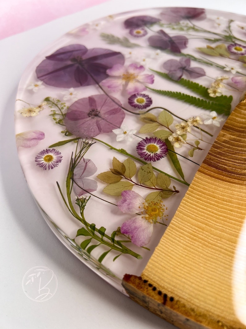 May include: A round, clear resin coaster with pressed flowers and leaves. The coaster has a light pink background and features purple, white, and green flowers. The coaster is attached to a wooden base.