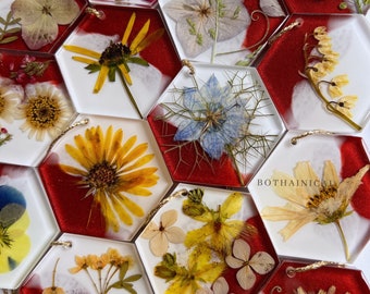 May include: Assortment of hexagonal resin ornaments with pressed flowers and leaves. Each ornament features a different floral design and is accented with a red and white background. The ornaments are strung with gold cord and have the word "BOTHAINICAL" printed on one.