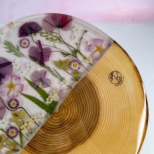 May include: A round wooden tray with a half-circle resin inlay featuring pressed flowers and leaves. The resin is clear and the flowers are purple, pink, and white. The wood is a light brown color and has a natural wood grain.