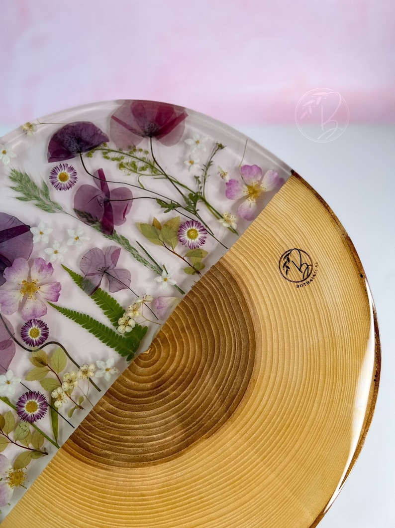 May include: A round wooden tray with a half-circle resin inlay featuring pressed flowers and leaves. The resin is clear and the flowers are purple, pink, and white. The wood is a light brown color and has a natural wood grain.