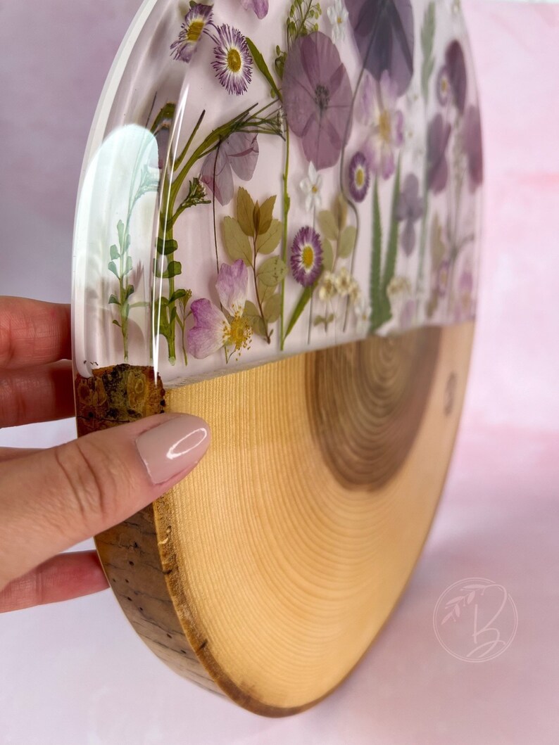 May include: A round wooden slice with a half-circle of clear resin containing pressed purple and white flowers. The resin is set on top of the wood slice, which has a natural wood grain.