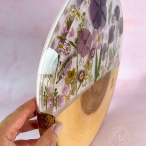 May include: A round wooden slice with a clear resin top, featuring pressed purple and white flowers. The resin is clear and shows the flowers in detail. The wood is light brown and has a natural grain.