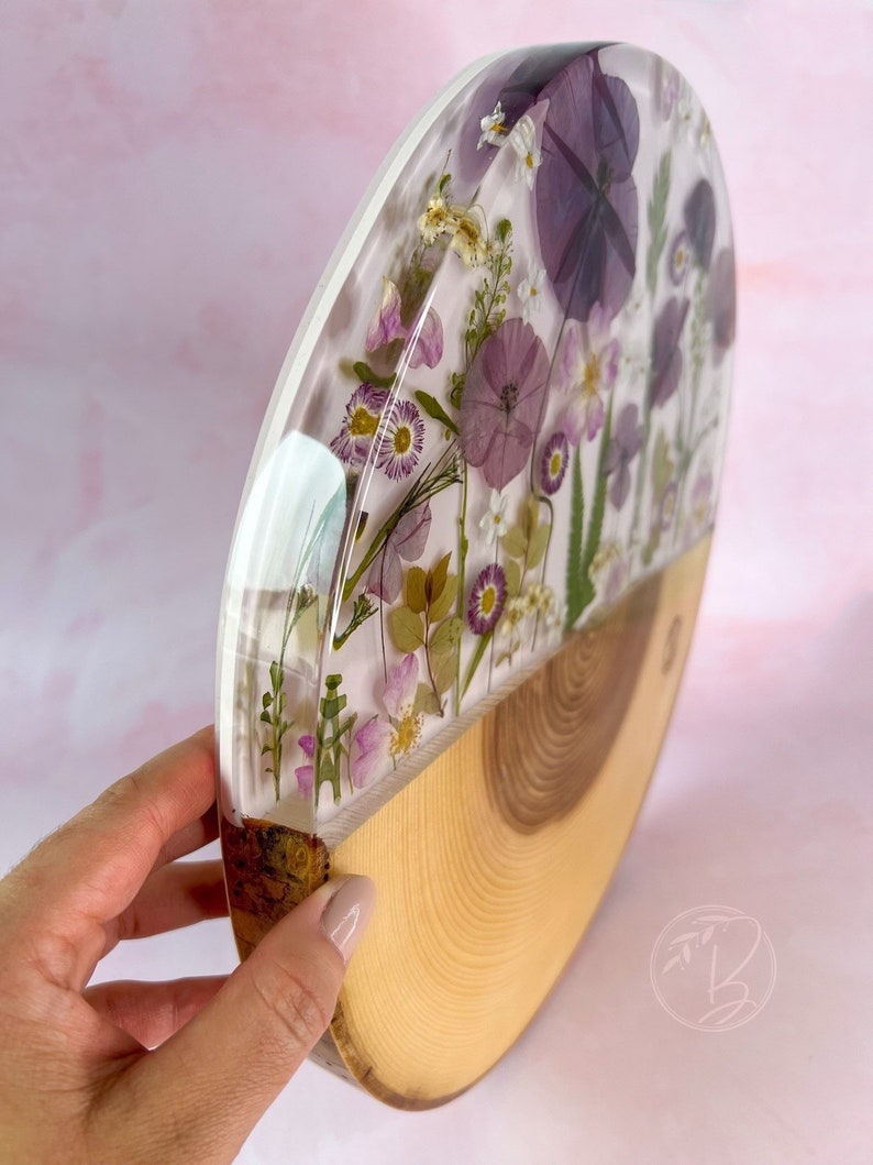 May include: A round wooden slice with a clear resin top, featuring pressed purple and white flowers. The resin is clear and shows the flowers in detail. The wood is light brown and has a natural grain.