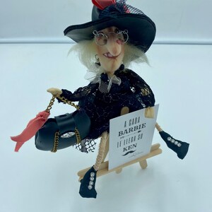 以下が含まれることがあります： 黒い帽子、赤いリボン、眼鏡をつけた黒い魔女の人形が、木製のイーゼルに座っています。人形は、金色のチェーンとピンクのリボンが付いた黒いハンドバッグを持っています。イーゼルには、「A cada Barbie le llega su Ken」と書かれた看板があります。