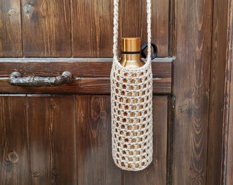Puede incluir: Un porta botellas de crochet beige con una botella de agua de cobre en su interior. El porta botellas está colgado de una puerta de madera.