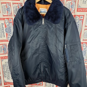 May include: Navy blue bomber jacket with a faux fur collar. The jacket has a zipper closure, a small pocket on the left sleeve, and ribbed cuffs. The lining is orange. The jacket is hanging on a wooden hanger.