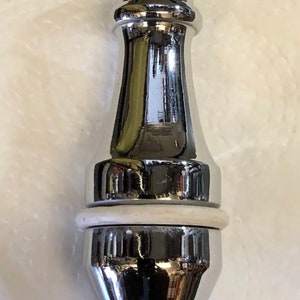 Puede incluir: Un tapón de botella decorativo cromado con un sello de goma blanco. El tapón tiene una forma redondeada y bulbosa con un cuello estrecho y una parte superior plana.