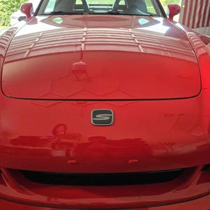 May include: A bright red sports car front view featuring a prominent hood, a black emblem with a silver S logo centered on the nose, side mirrors, and a lower air intake grille. The car is parked in a garage with a flag visible in the background.