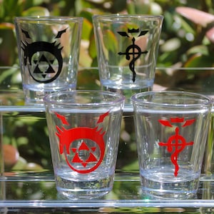 May include: Four clear shot glasses with black and red graphic designs. Two glasses have a black design, and two have a red design. The shot glasses are displayed on a clear glass shelf, with a blurred green background.