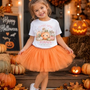 May include: A white short-sleeved t-shirt featuring a graphic of pumpkins and the text Cutest Little Pumpkin In The Patch, paired with an orange tulle skirt and white sneakers. The shirt design includes various pumpkins in soft autumn colors.