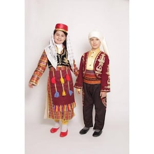 Puede incluir: Dos niños vistiendo ropa tradicional turca. La niña lleva un vestido rojo y dorado con un pañuelo blanco en la cabeza y el niño lleva una chaqueta roja y dorada con un sombrero blanco.