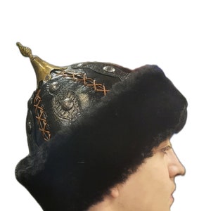 May include: A black leather hat with a fur trim and a gold metal ornament on top. The hat has a decorative design on the front.