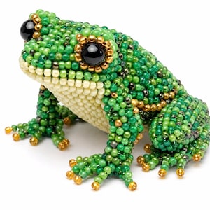 May include: A decorative frog figurine crafted from small, multi-tonal green and gold glass beads. The frog features large, glossy black bead eyes surrounded by metallic gold rings, with a cream-colored beaded underbelly and textured skin.